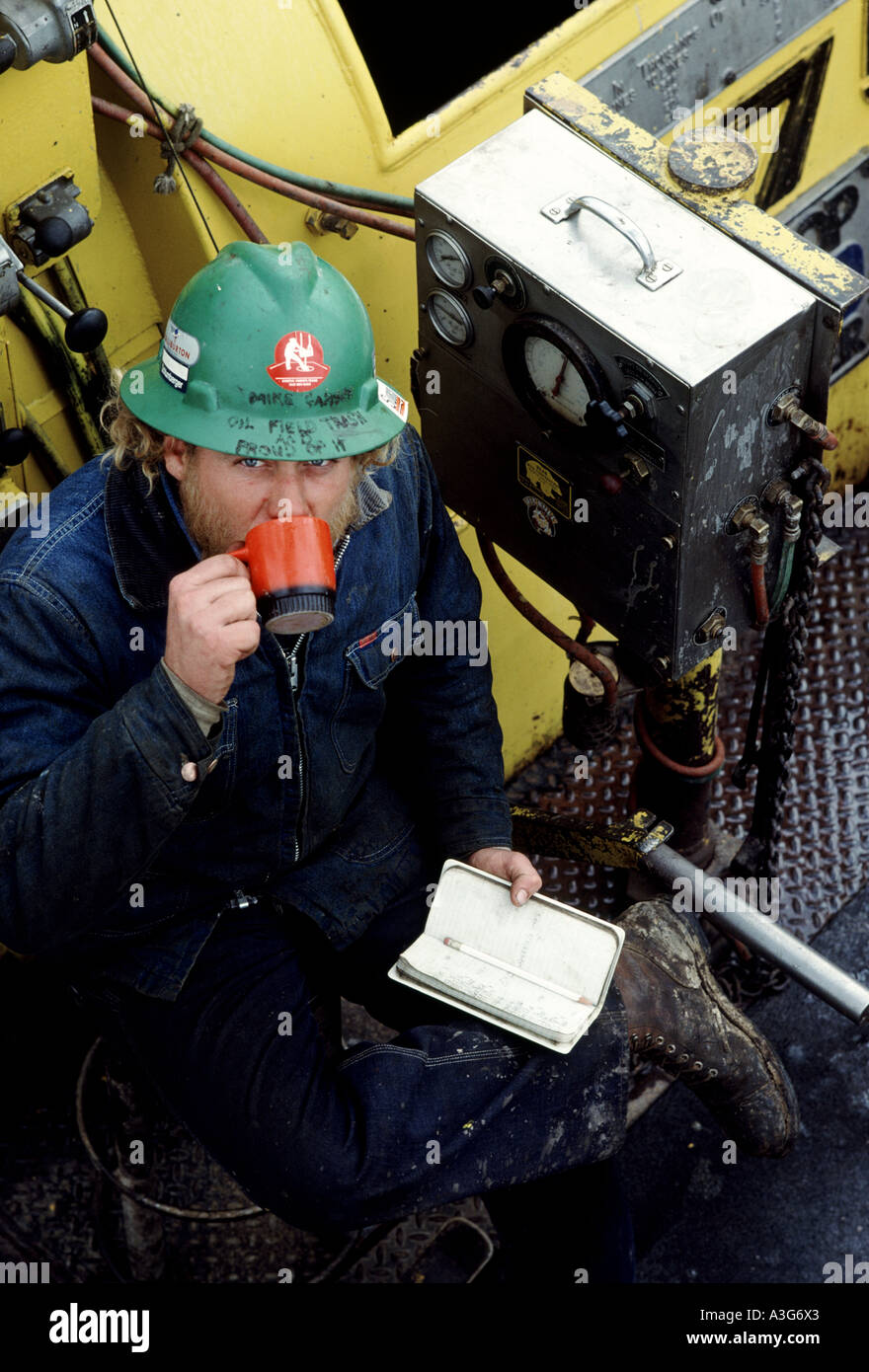 Overhead view of oilman in a green hardhat drinking a hot beverage, sitting to plan his day on an off shore drilling platform. Stock Photo