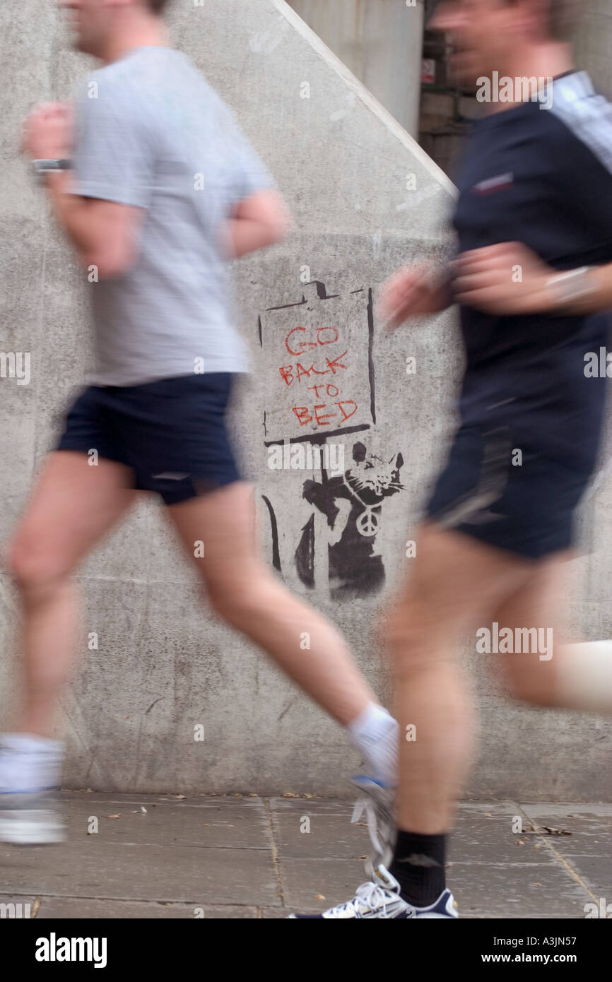 Joggers running past Banksy stencil graffiti of rat holding Go Back To Bed sign. Thames Embankment, London, England Stock Photo