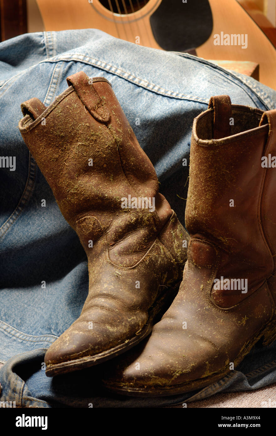 Cowboy boots covered in dried hay and grass and worn out leather against an old denim jacket with guitar Canada Stock Photo