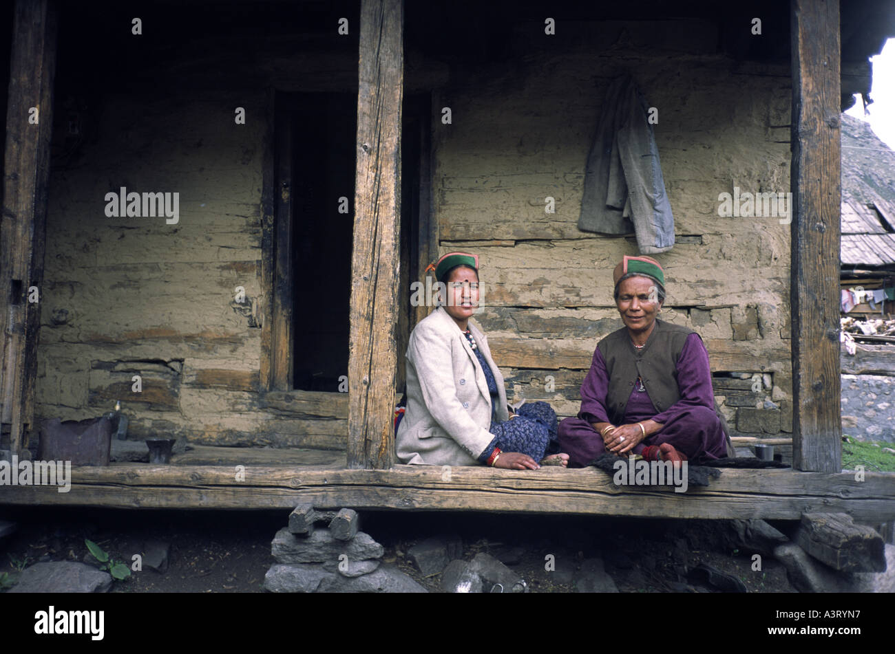 Two local women in traditional dress and wearing Thepang hats, sit outside a wooden house in Chitkul, Himachal Pradesh India Stock Photo