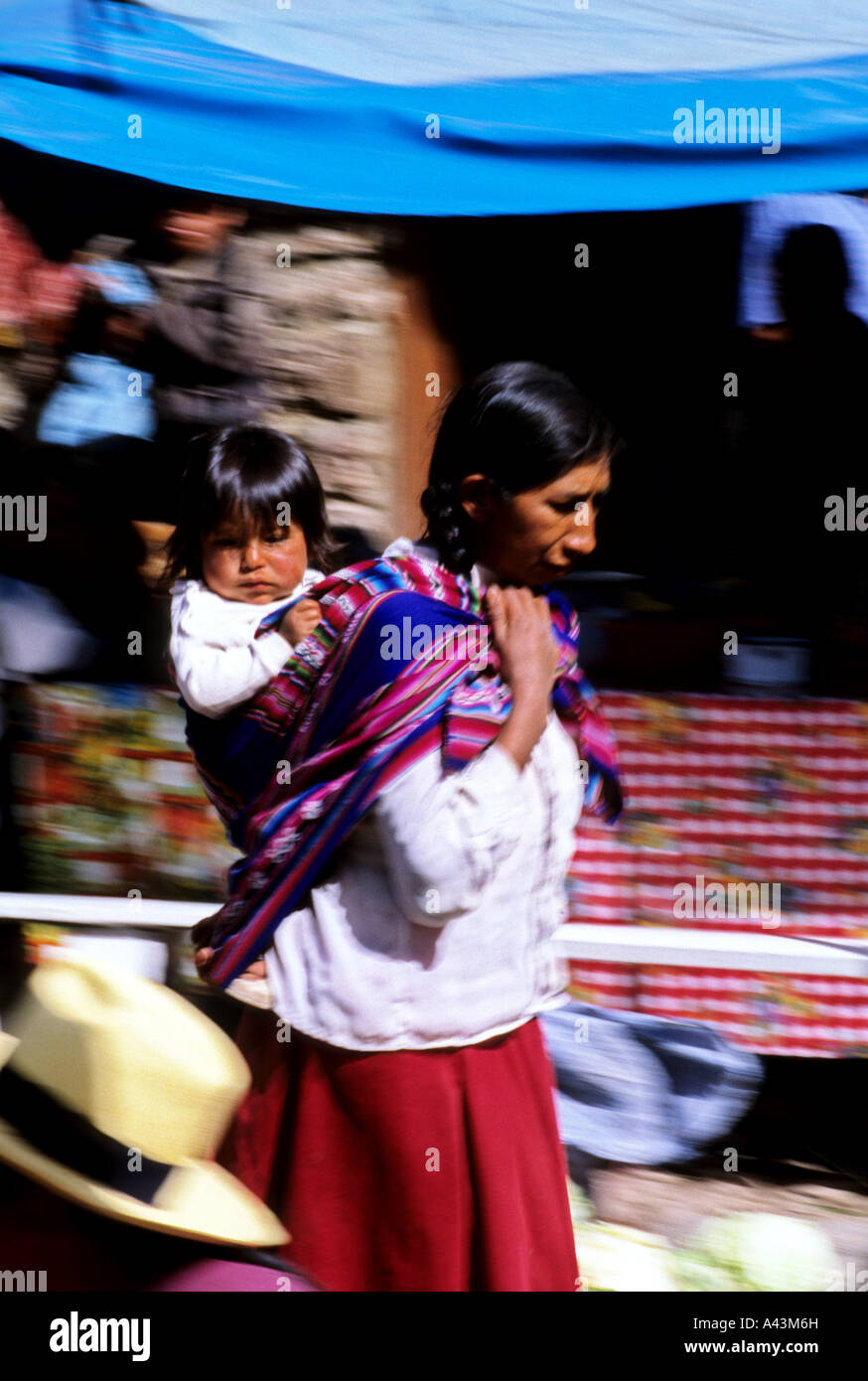 Traditionally dressed woman carrying child on back through the market in the Incan village of Pisac Sacred Valley Peru Stock Photo
