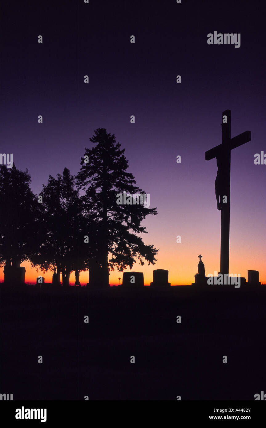 A crucifix and evergreen trees stand silhouetted at sunrise in an unknown cemetery on the Minnesota plains Stock Photo