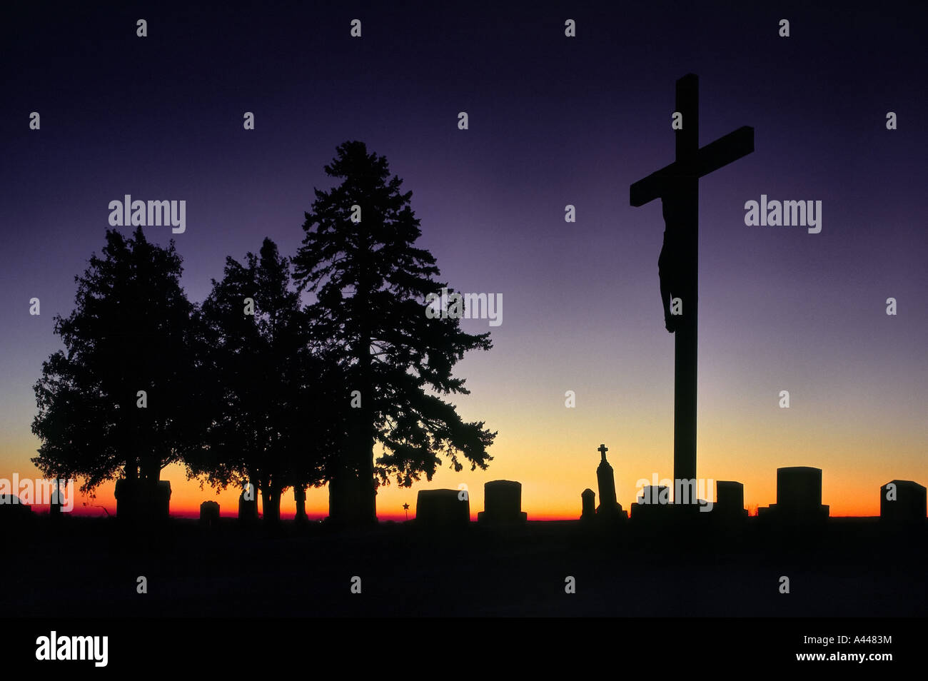 A crucifix and evergreen trees stand silhouetted at sunrise in an unknown cemetery on the Minnesota plains Stock Photo