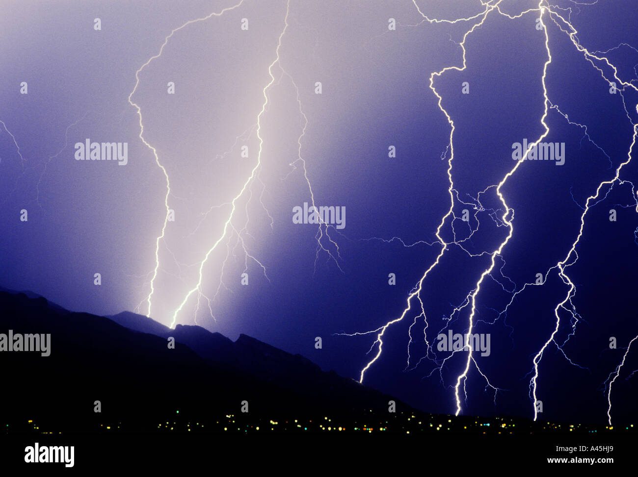 Bolts of lightning strike against a blue sky over a mountain range and city lights in Southern Arizona, USA. Stock Photo