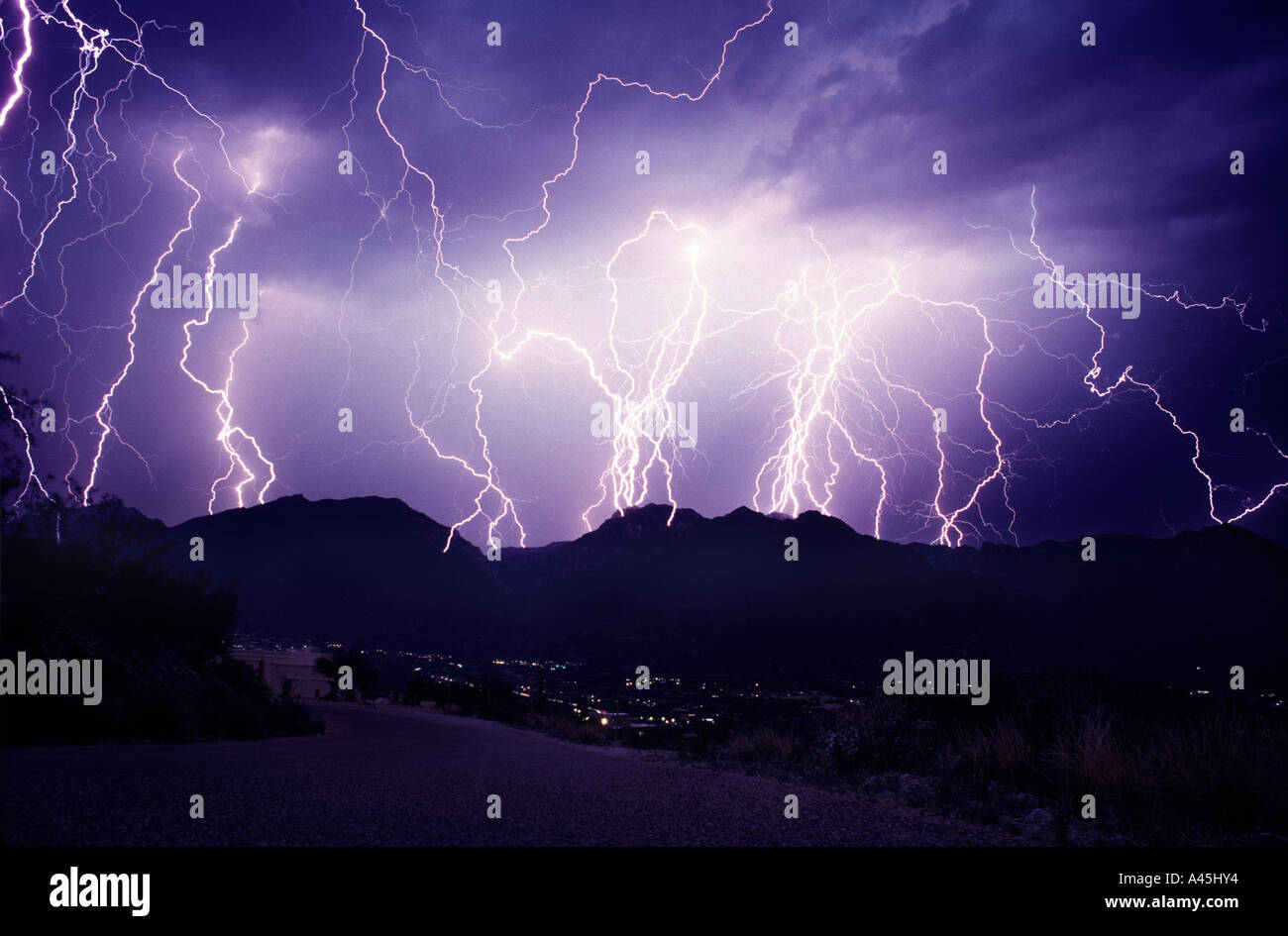 Lightning strikes over a city dotted with glowing lights from a dark blue stormy sky in Southern Arizona during a monsoon storm. Stock Photo