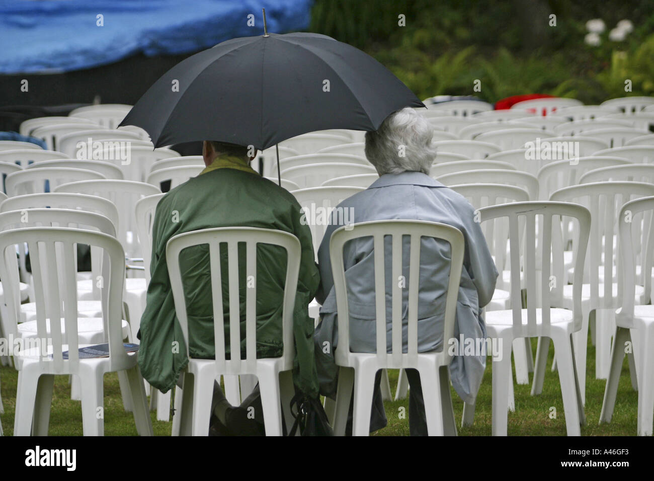 Two elderly English old age Pensioners sit on chairs in the rain waiting for an open air opera. Stock Photo