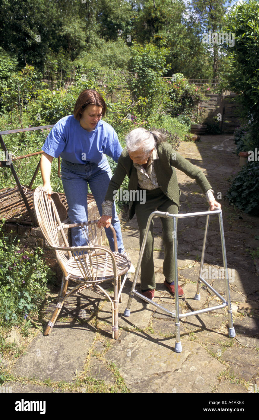 carer helping 84 year old woman on walker sitting on garden chair at home Stock Photo