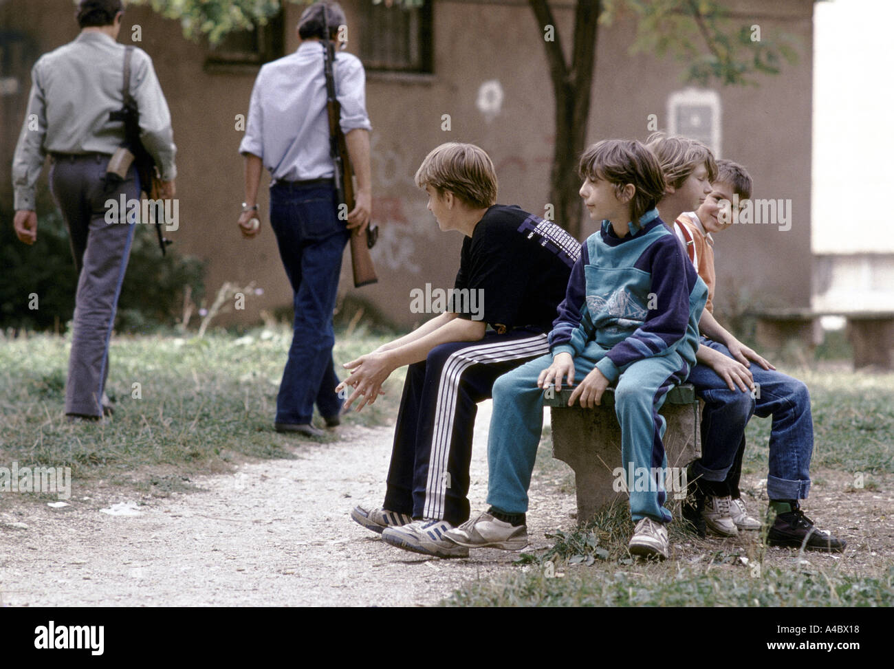 Men with machine guns walking past group of children sitting on a public bench in the Serb-controlled area of Grbavica, Sarajevo Stock Photo