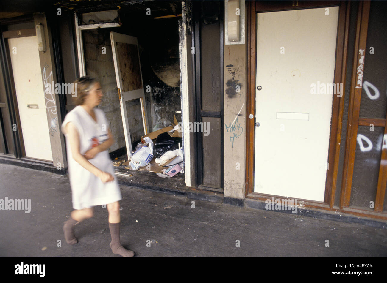 A WOMAN IN PYJAMAS & SOCKS WALKING PASS A DOORWAY OF A DERELICT FLAT LITTERED WITH GARBAGE IN A HOUSING ESTATE Stock Photo