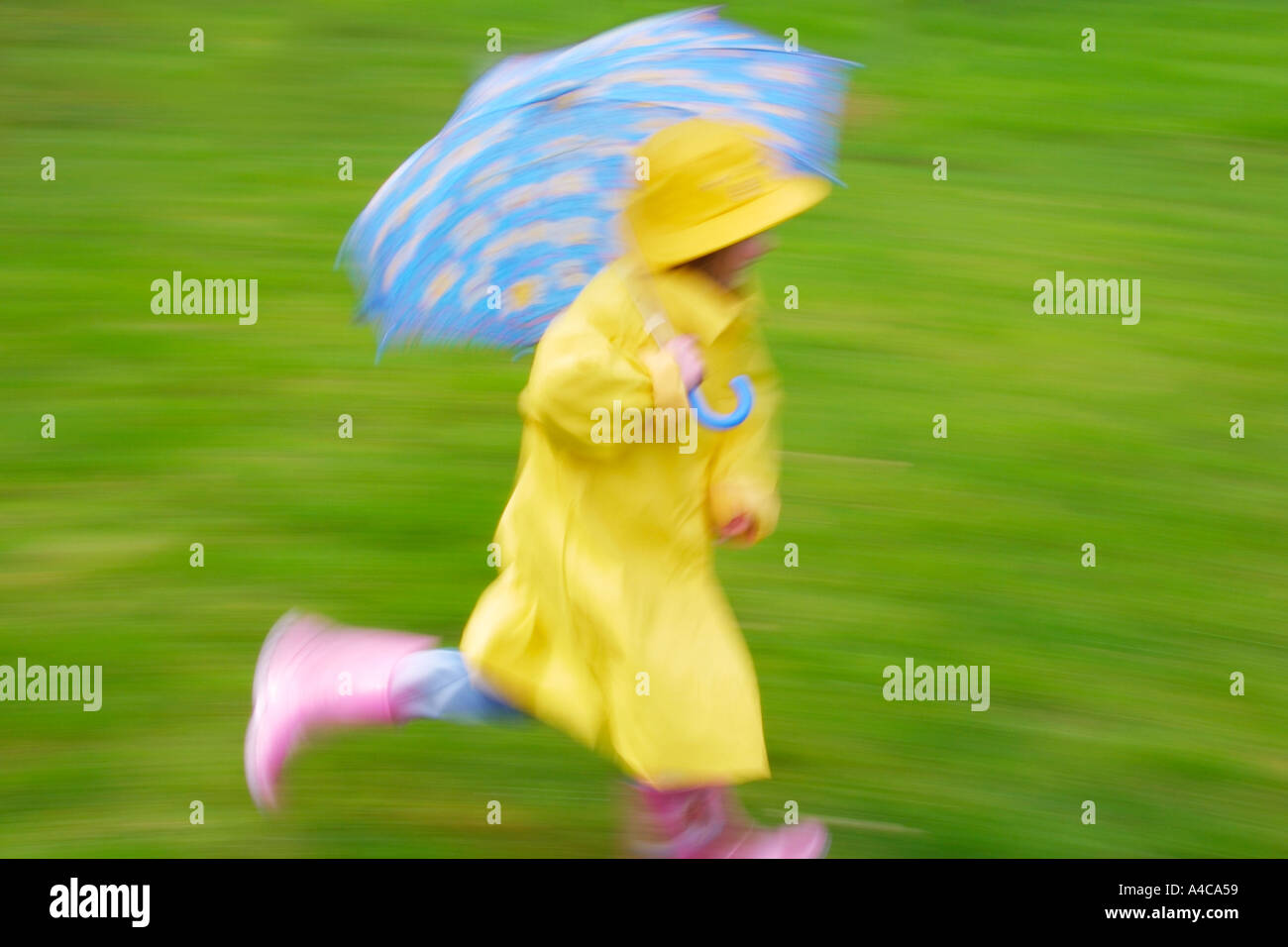 YOUNG GIRL RUNNING WITH BLUE UMBRELLA YELLOW RAINCOAT AND SOU'WESTER AND PINK WELLINGTON BOOTS  BLURRED IMAGE UK Stock Photo
