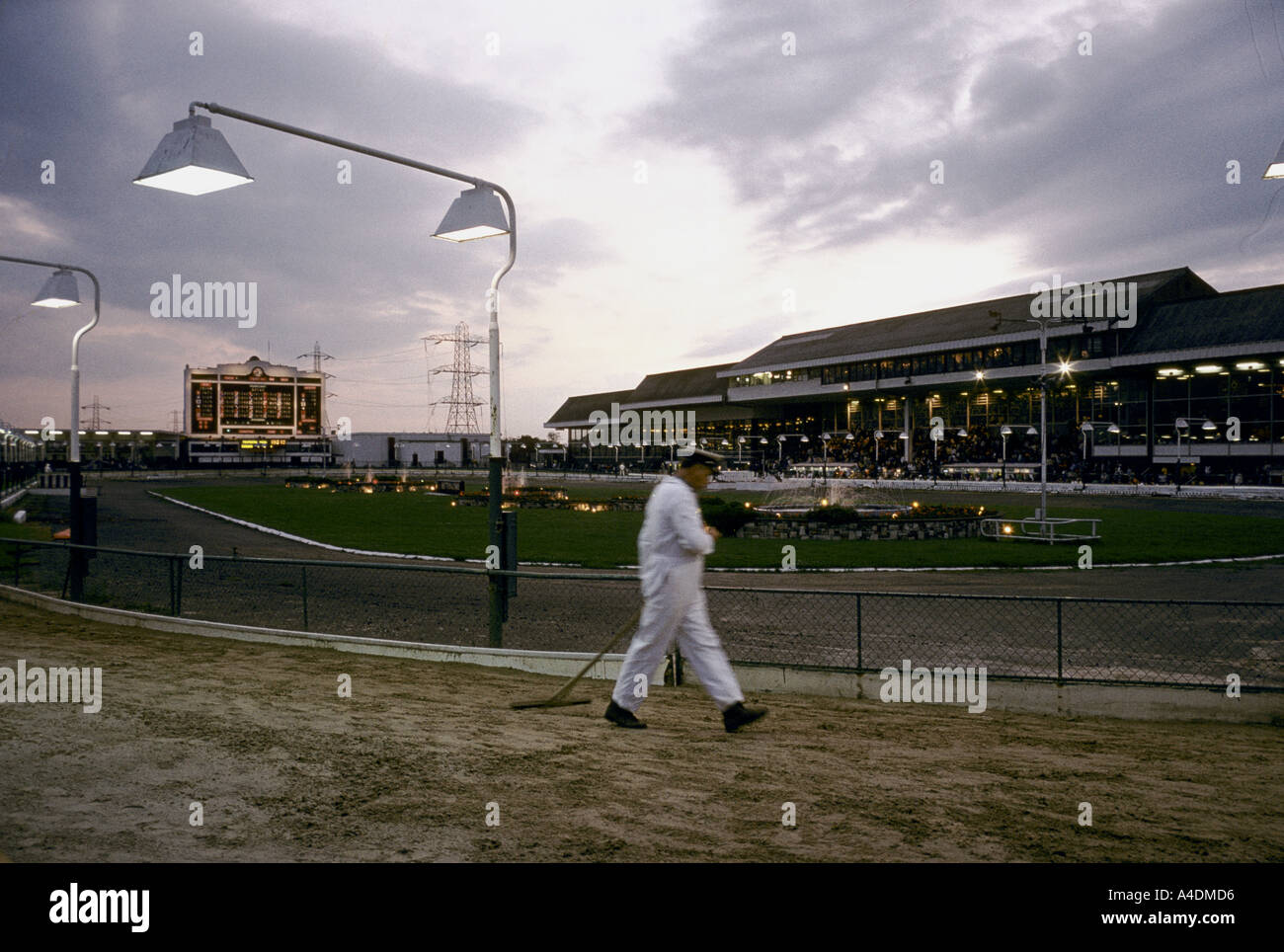 A male assistant wearing a white boiler suit raking the track for greyhound racing as the sun sets over Walthamstow Stadium Stock Photo