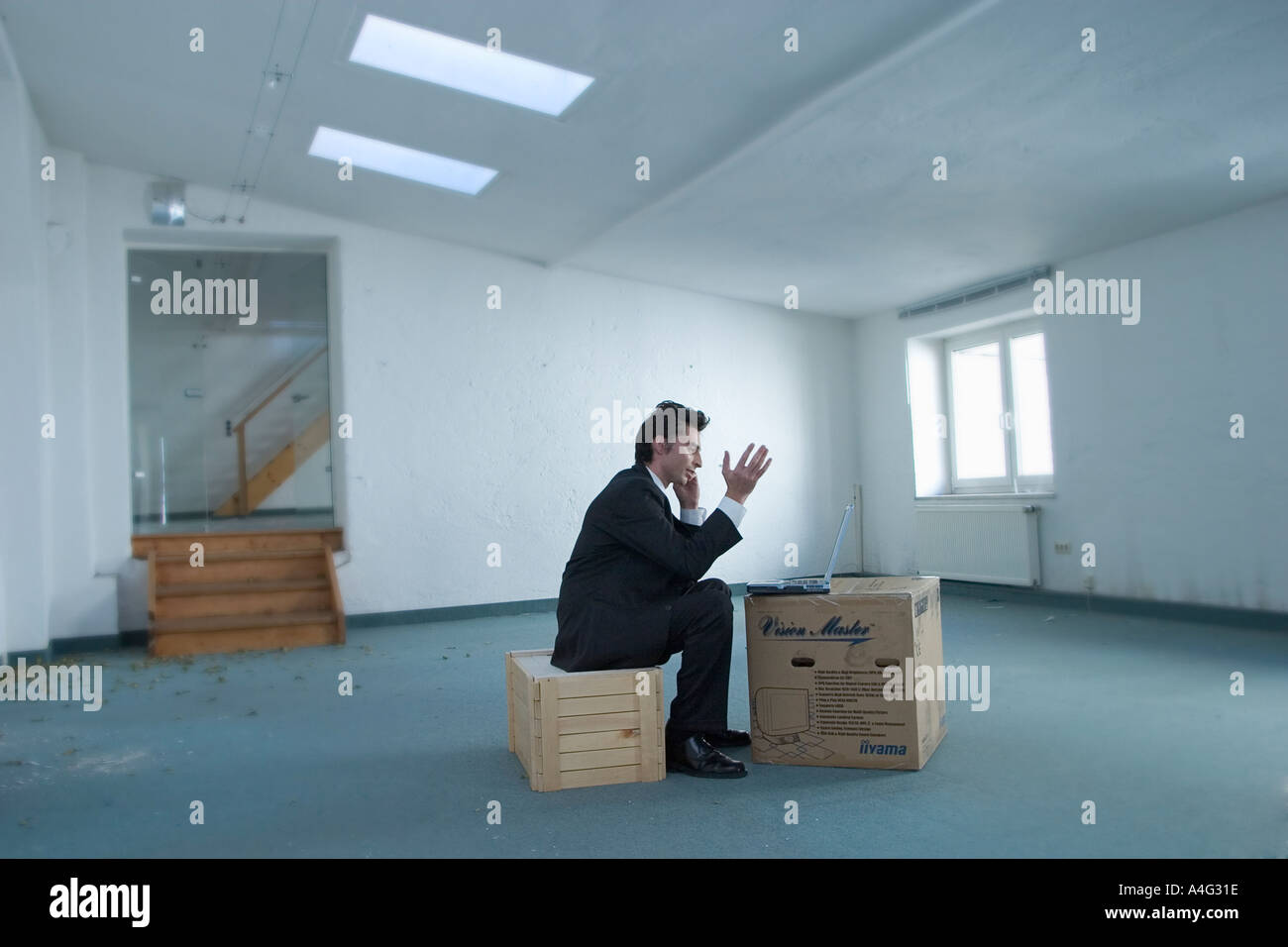 MR Man in business outfit sits with his laptop on a box in an empty loft and telephones Stock Photo