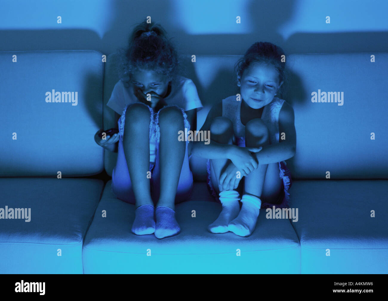 Two little girls sitting on sofa watching television in dark, laughing Stock Photo
