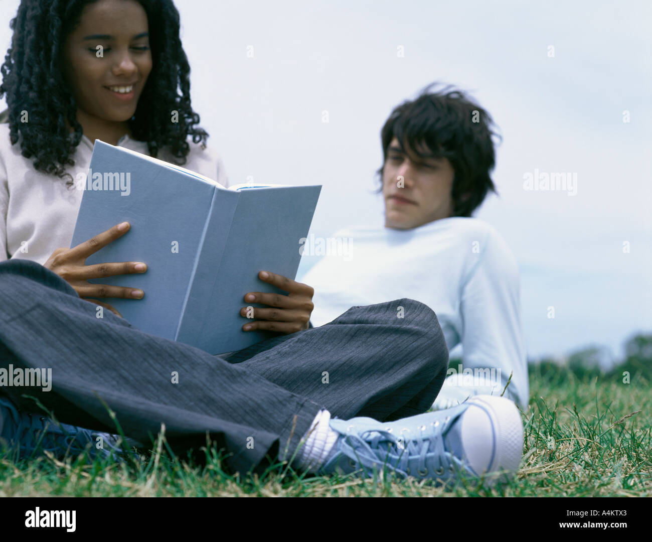 Young woman sitting on grass reading book, young man leaning on elbow behind her Stock Photo