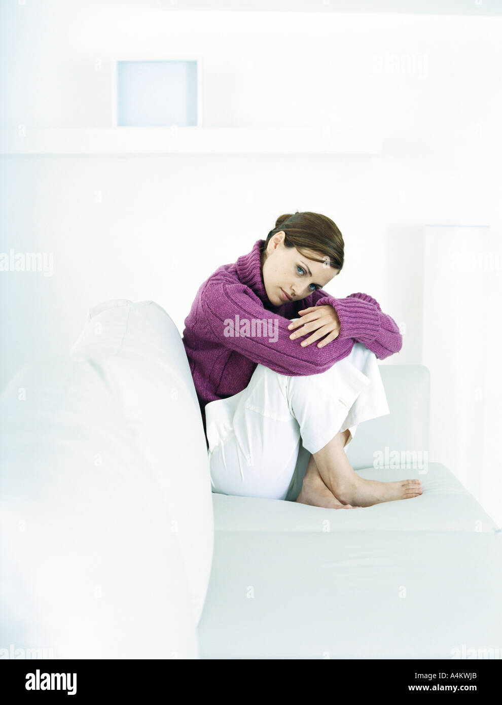 Young woman sitting barefoot on sofa with knees up and head bent forward Stock Photo