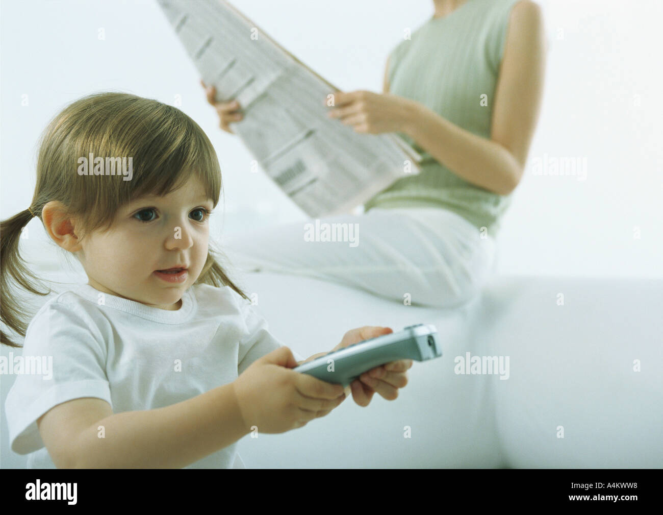 Little girl pointing remote control, woman sitting on back of sofa holding newspaper in background Stock Photo