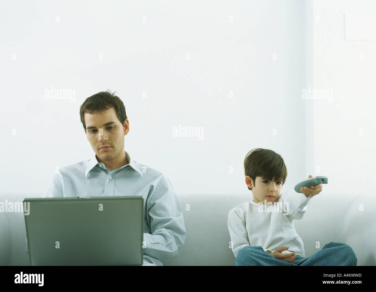 Man sitting on sofa working on laptop, boy sitting next to him pointing remote control Stock Photo