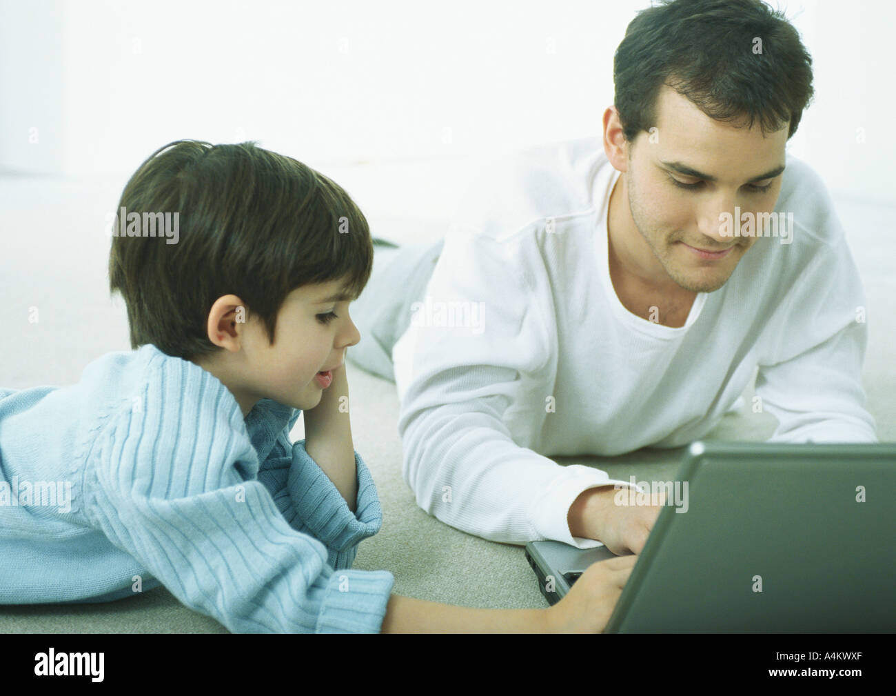 Man and little boy lying on floor on stomachs, working on laptop Stock