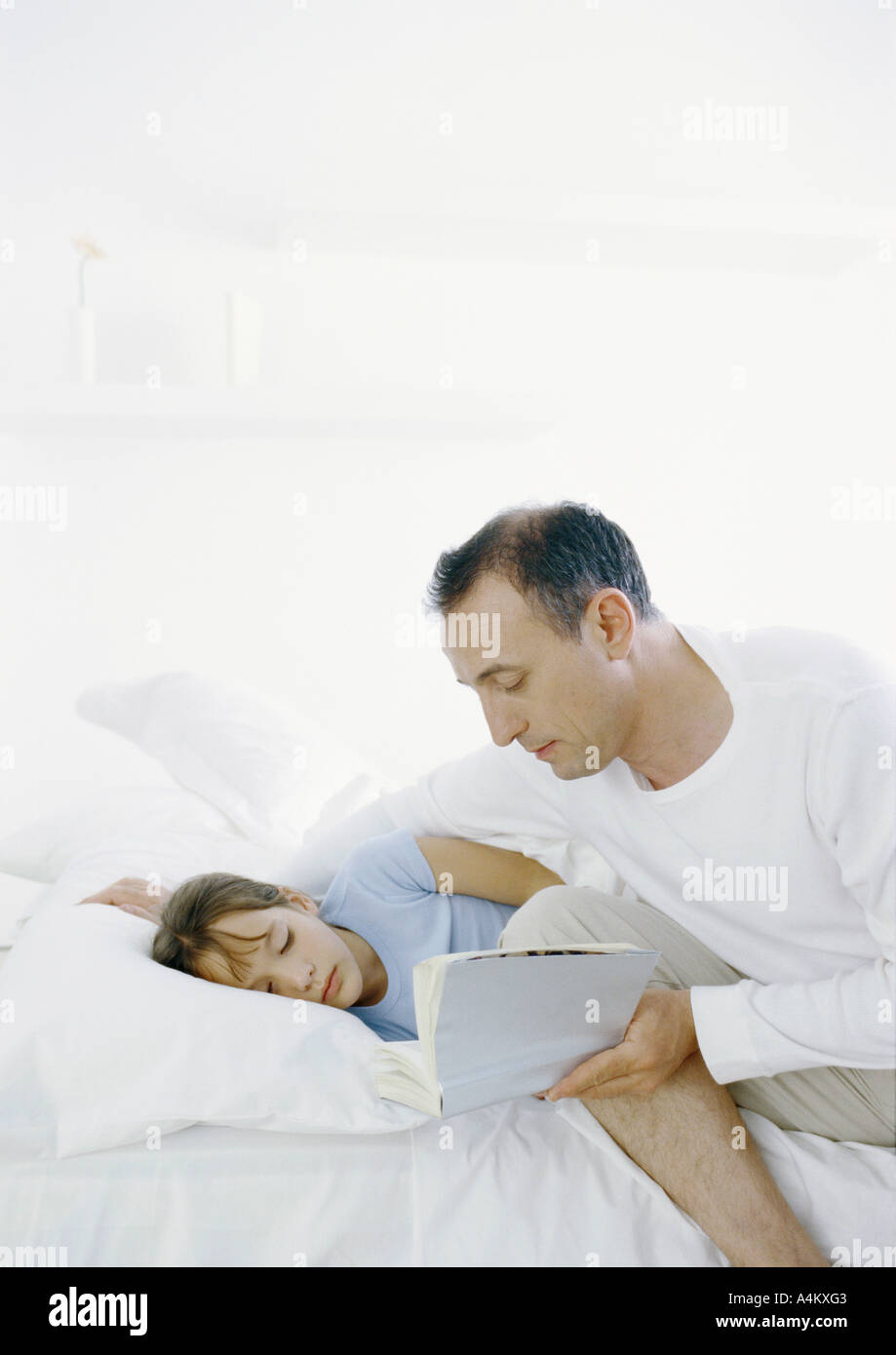 Man sitting on edge of bed reading book to girl lying in bed Stock