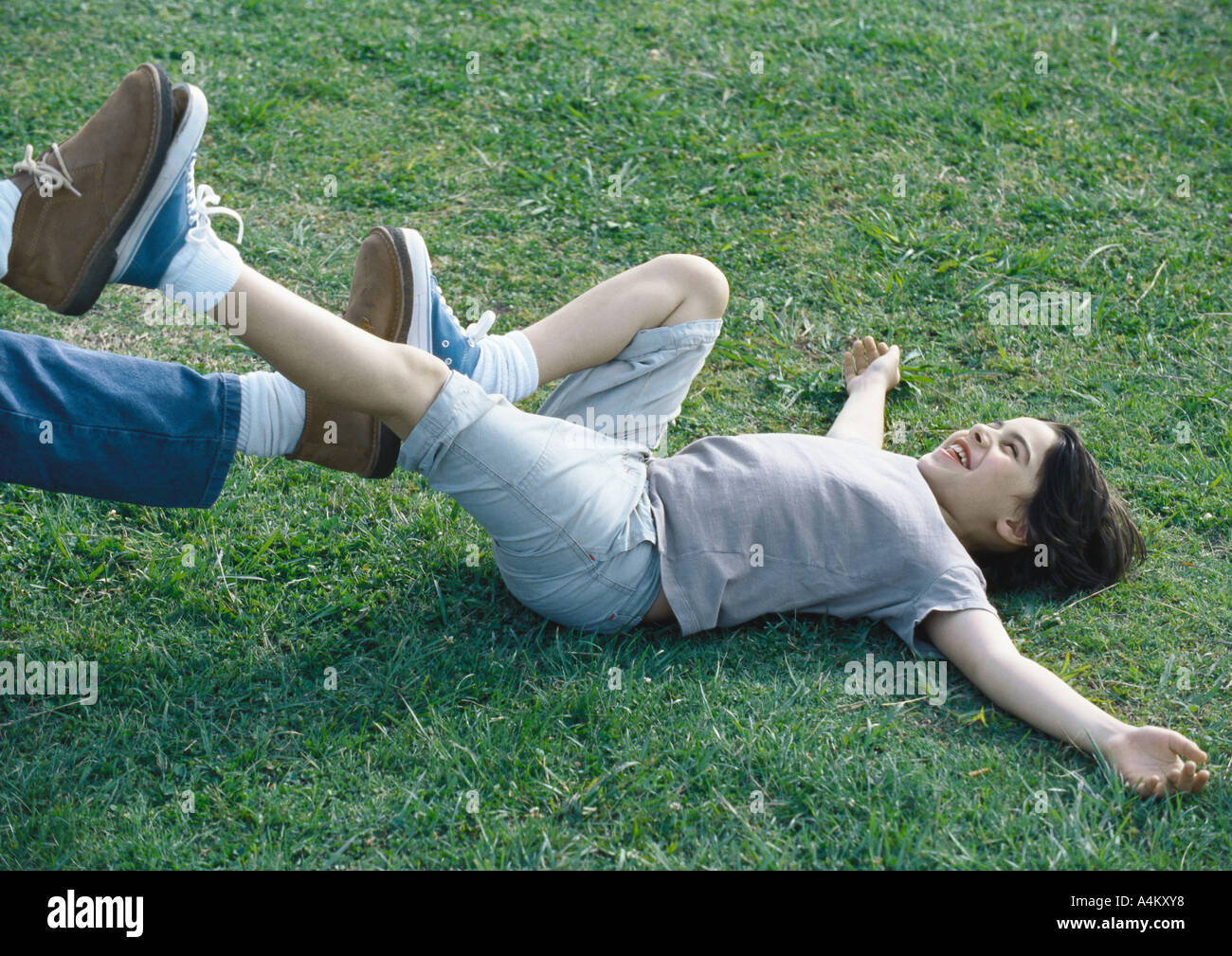Boy lying on back on grass with feet pushing against man's feet Stock