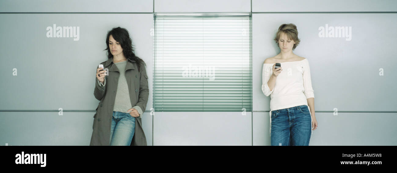 Two young women leaning against wall, looking at cell phones Stock Photo