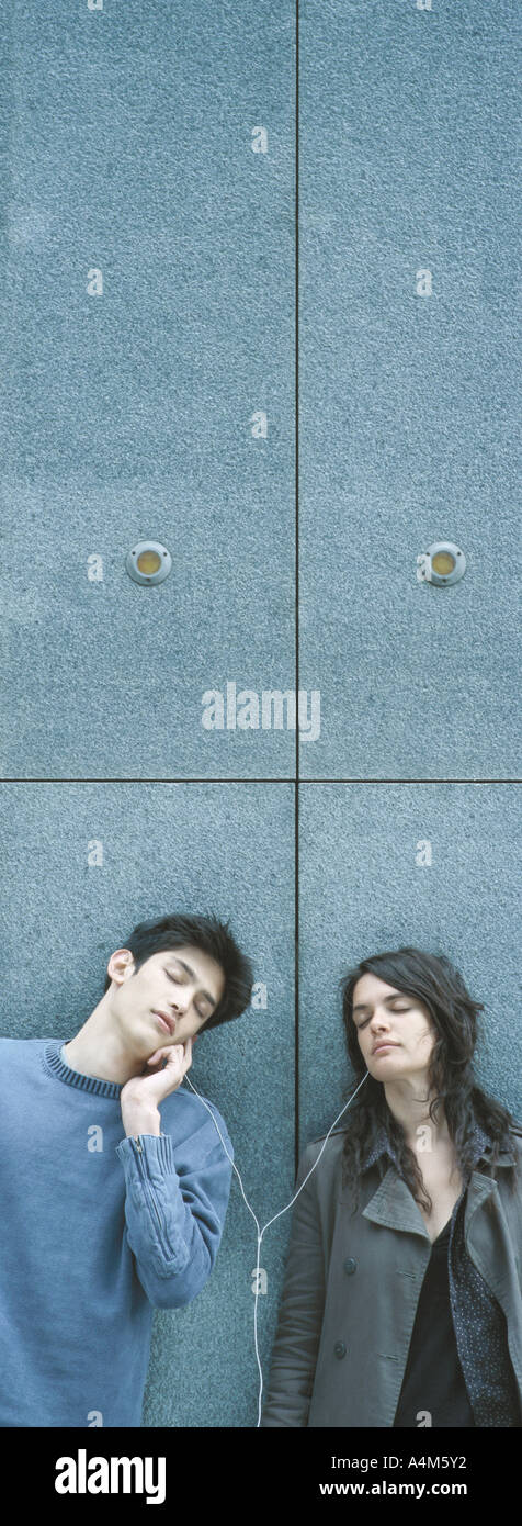 Young couple leaning against wall, sharing earphones, listening to music with eyes closed Stock Photo