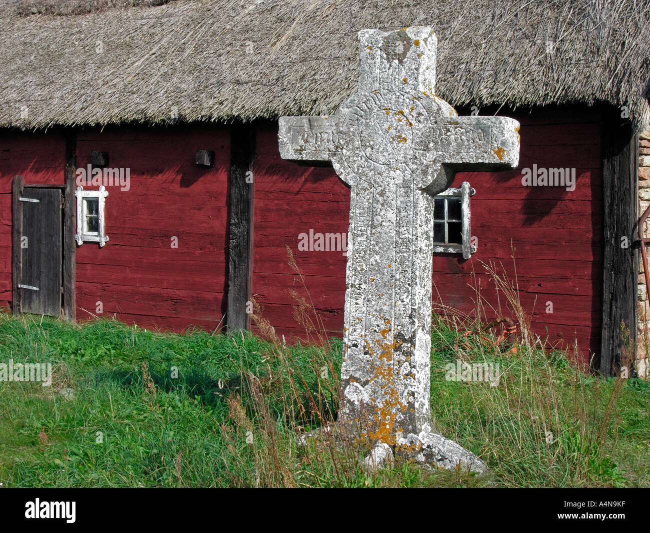 old granit stone cross in front of a red wooden house with thatched roof island Oland Stock Photo
