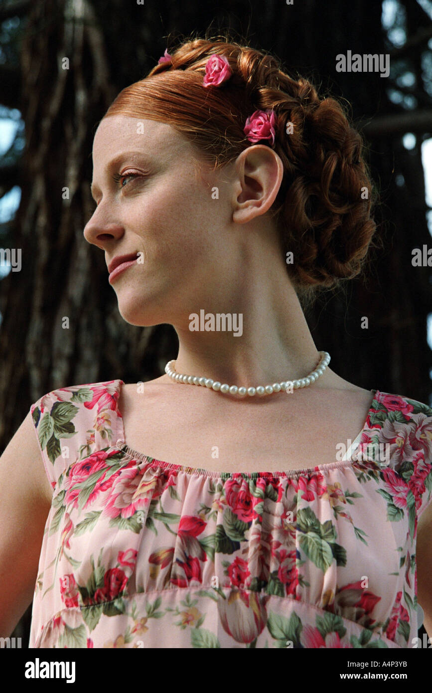 Young woman with red hair in a floral dress and pearl necklace in the trees with flowers in her hair Stock Photo
