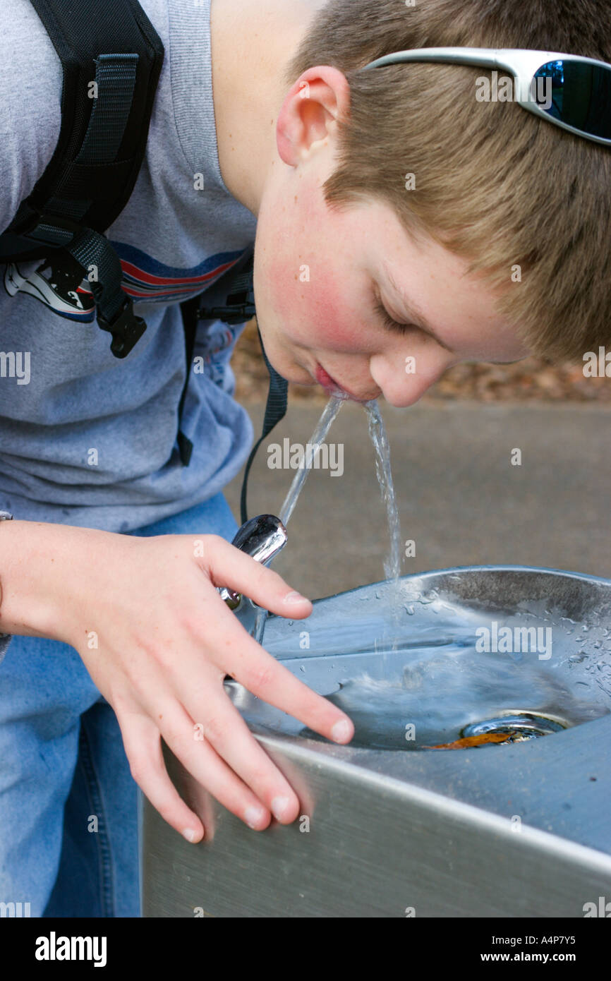 Male teenager with sunglasses on head and carrying a backpack drinking from a water fountain outdoors Stock Photo