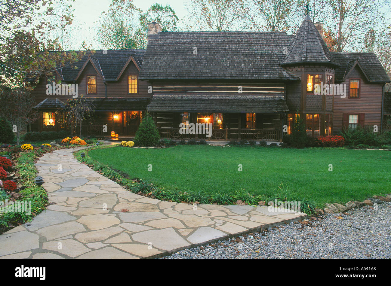 Front exterior large high-end historical log cabin style house with additions on each side -- lit up for fall night and a welcoming yard. USA, Stock Photo
