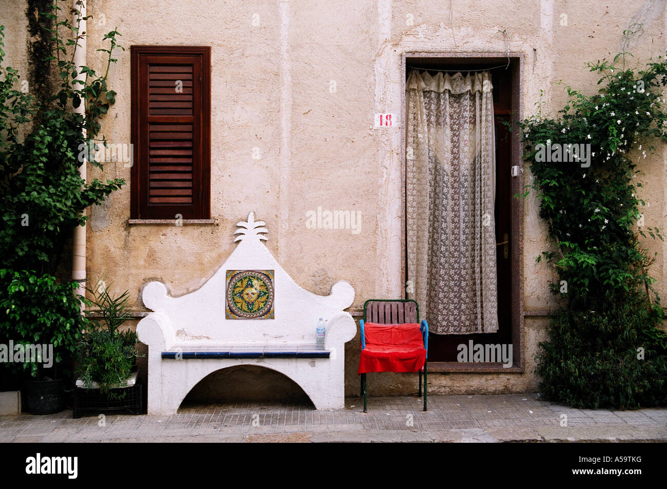 A view of a bench and chair outside the front of a home.  Net curtain in doorway. Wooden shutter. Climbing plant on the wall Stock Photo