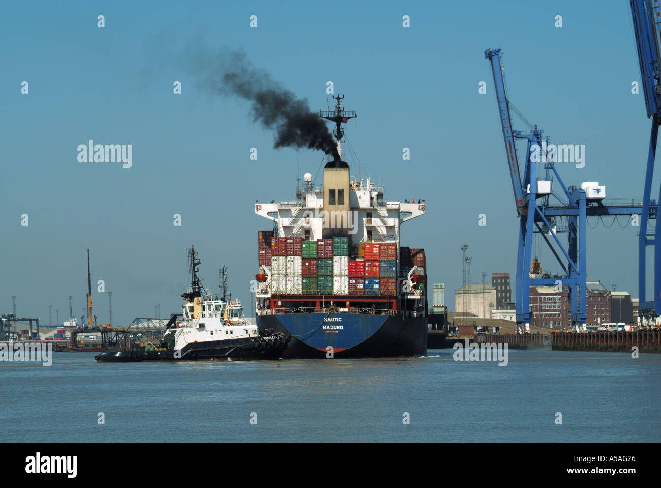 Arriving cargo container freighter ship & black smoke stacked with load shipping containers tugboat pushes towards cranes Felixstowe port Suffolk UK Stock Photo