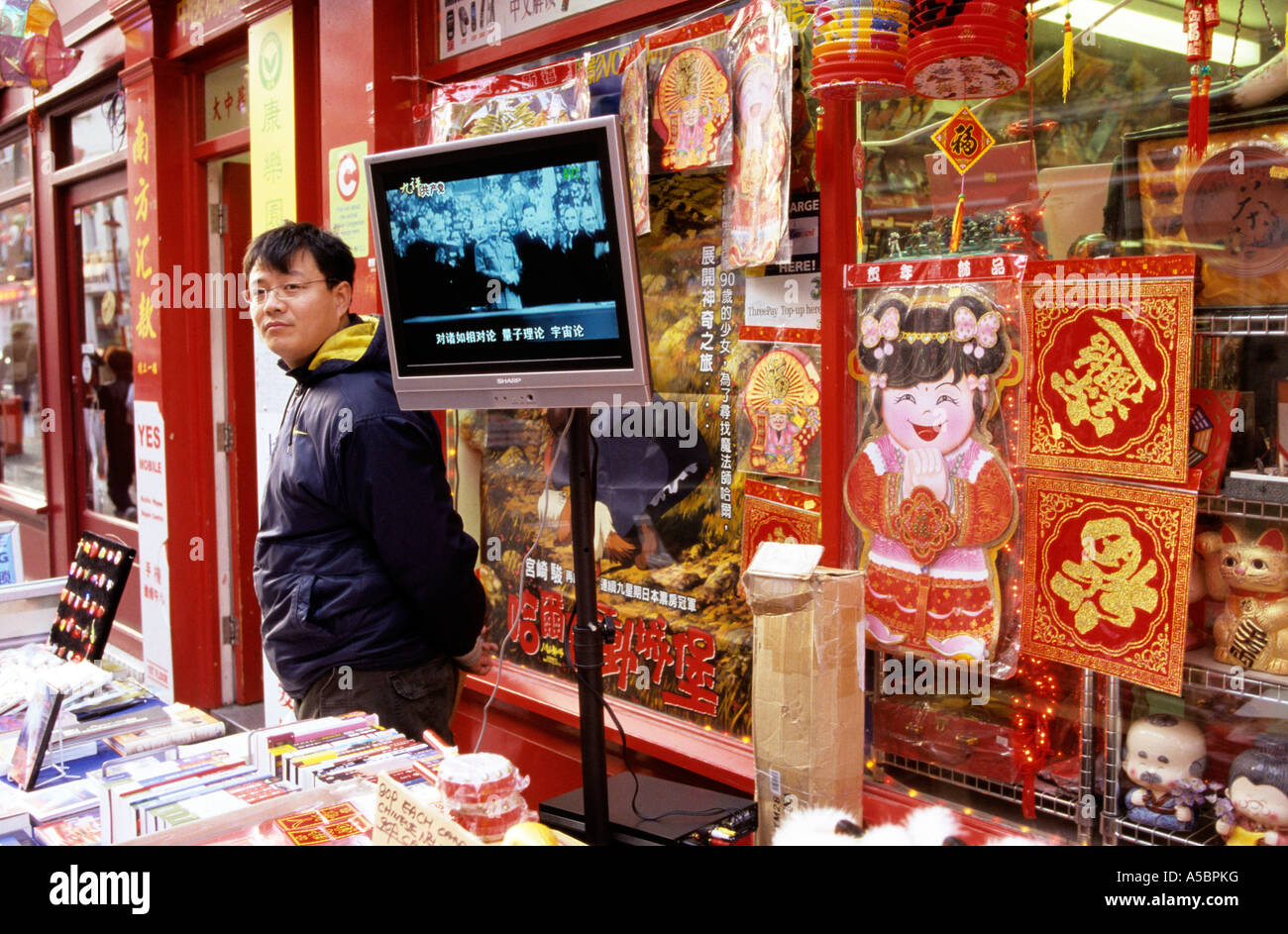 Chinese man manning stall, standing beside flat screen television, Chinatown, London, England, UK Stock Photo