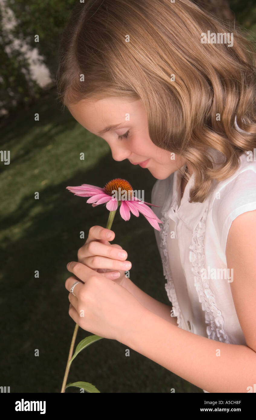 A color vertical image of a young blonde girl in a white blouse smelling a big pink flower with orange center near her face Stock Photo