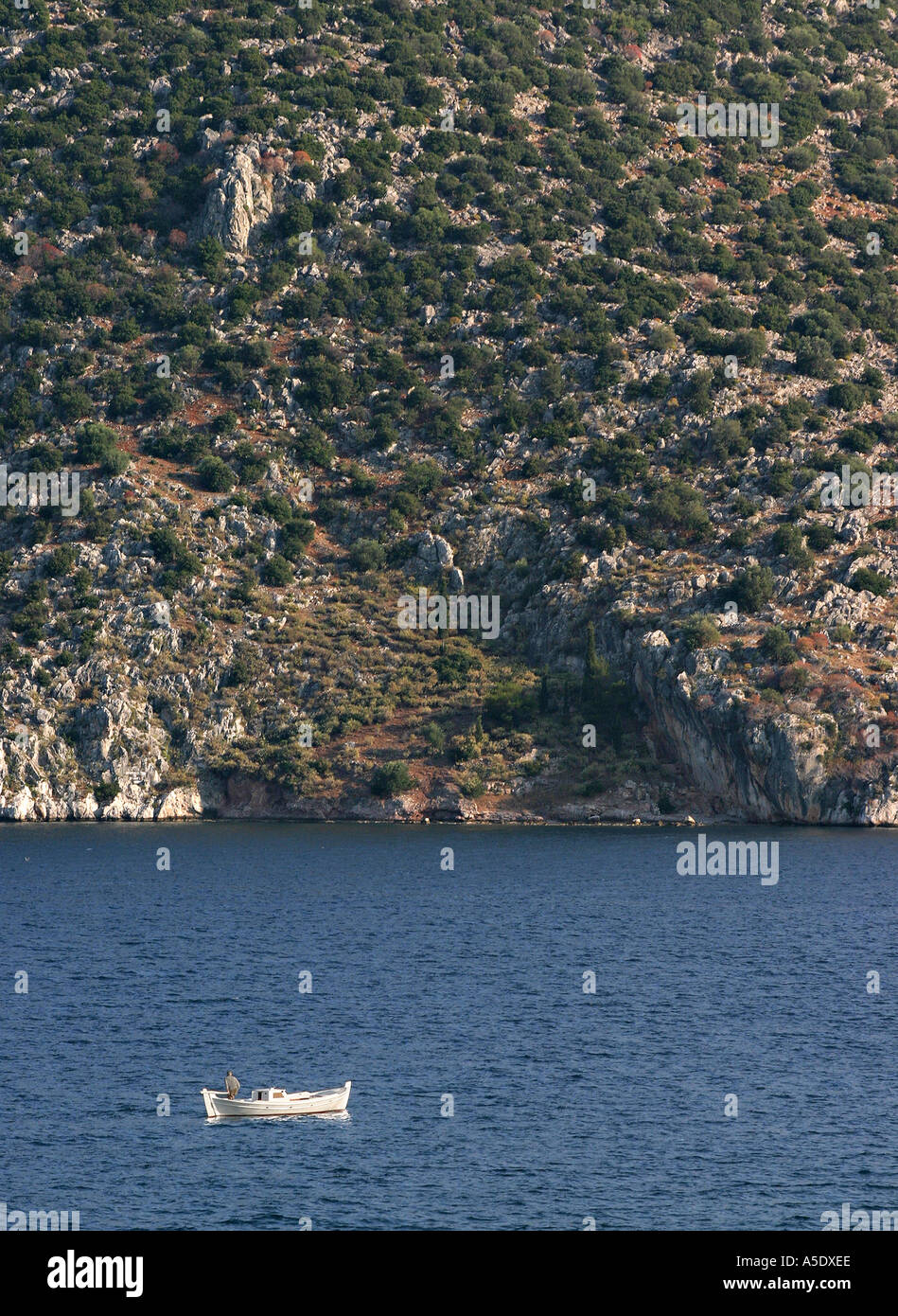 Lone fisherman in a white boat on blue waters against a scrubby hillside in the background Tolo Peloponese Greece Stock Photo