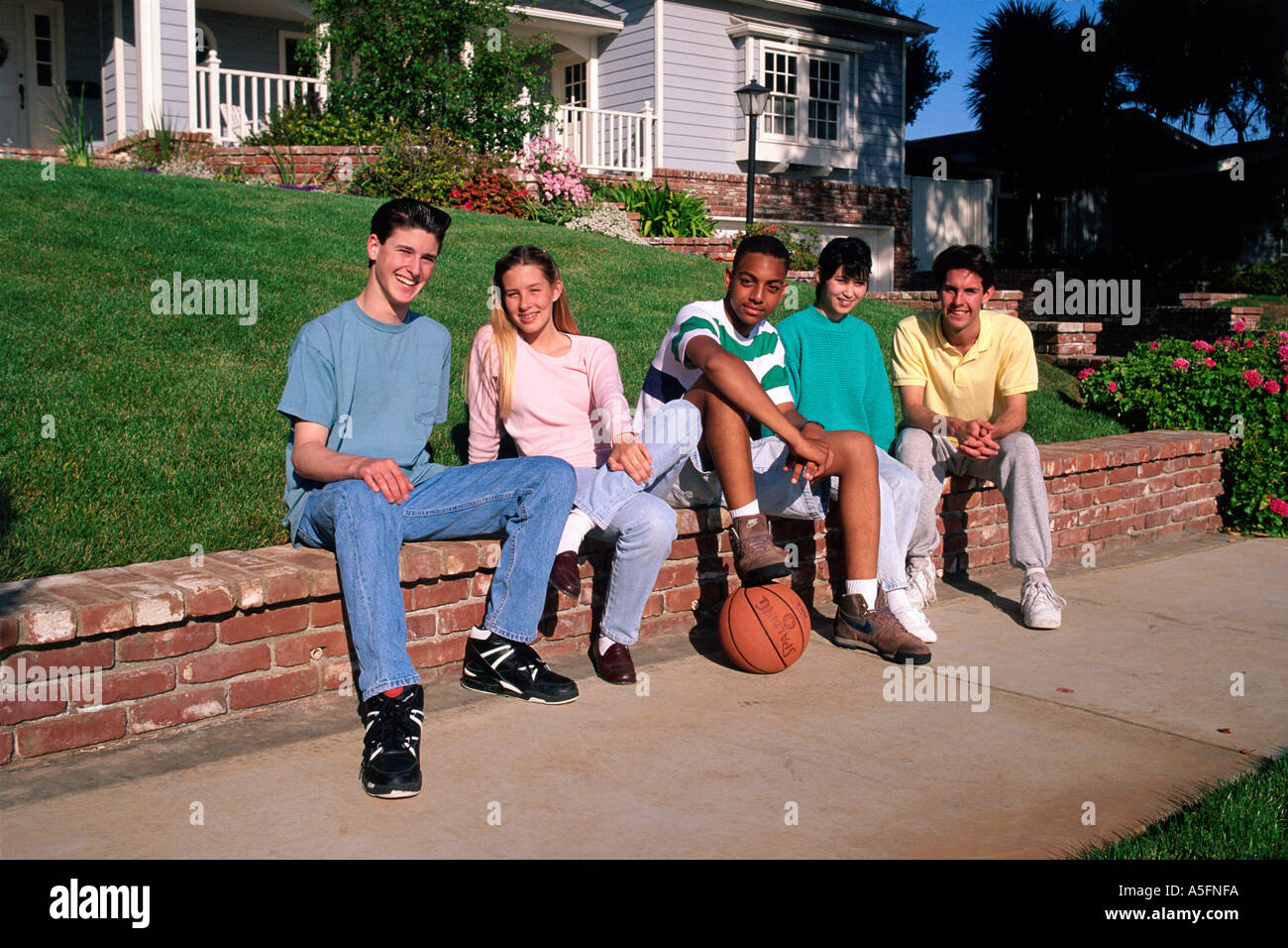 racially diverse, smiling, teenagers, friends sit on low wall near sidewalk, near home, with soccer ball Stock Photo
