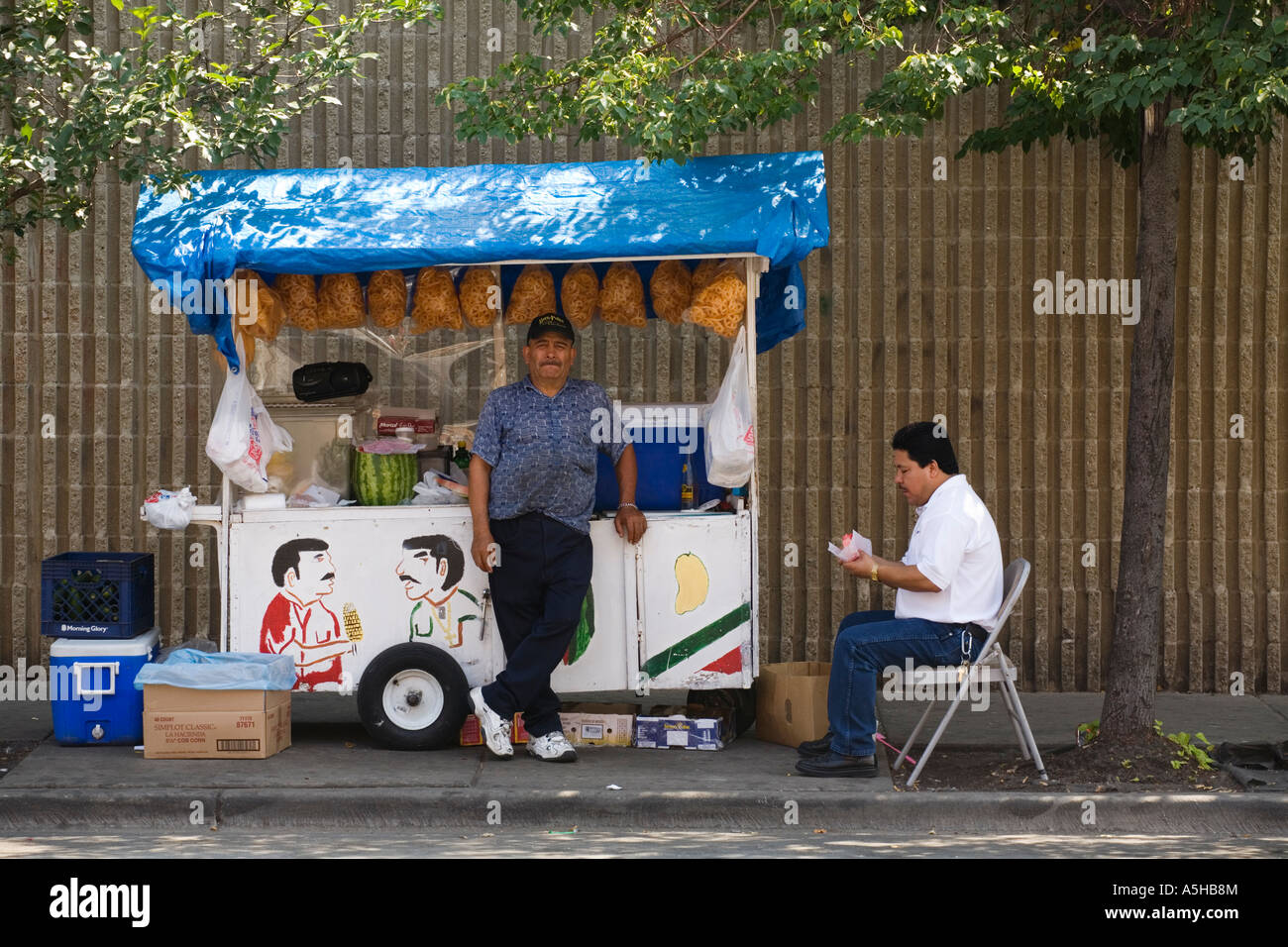 ILLINOIS Chicago Man sit in folding chair on sidewalk eat food purchased from street vendor man stand by cart sell food Stock Photo