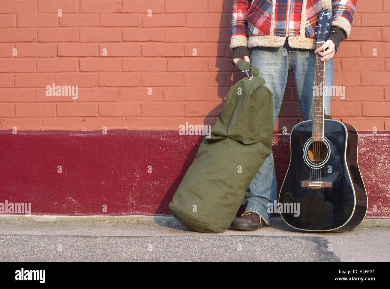 man stood against red wall with duffel bag and guitar Stock Photo