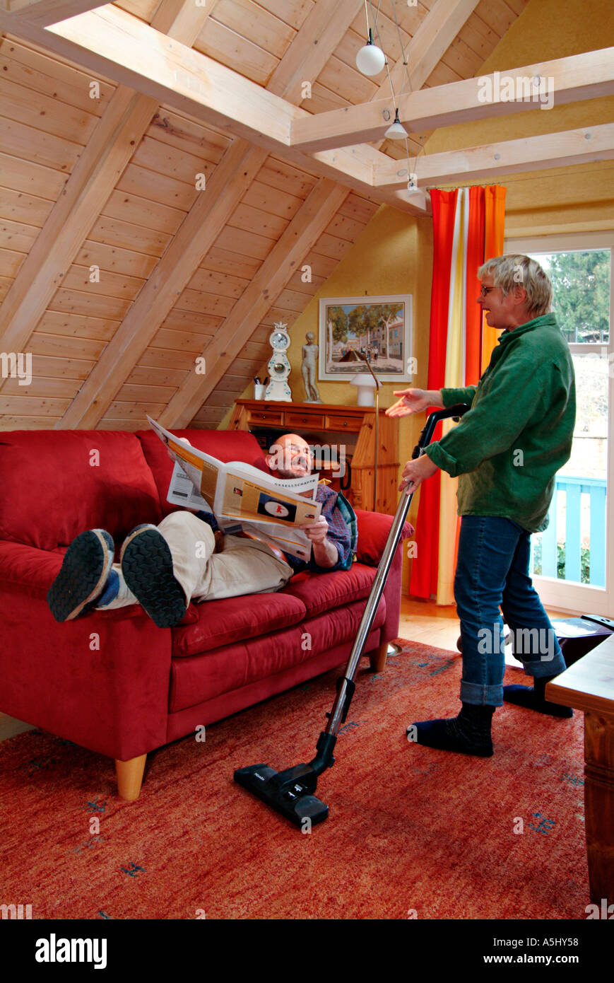 MR PR woman vacuuming hoovering and grousing with a man lying on a sofa reading a newspaper Stock Photo