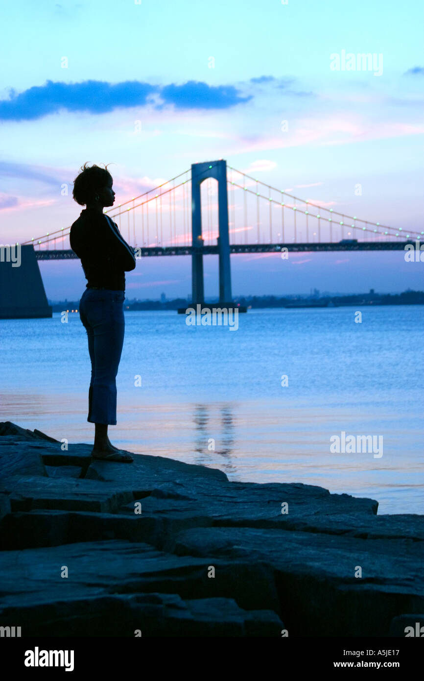 African American black female woman silhouetted standing on jetty rocks in front of Throgs Neck Bridge at sunset Stock Photo