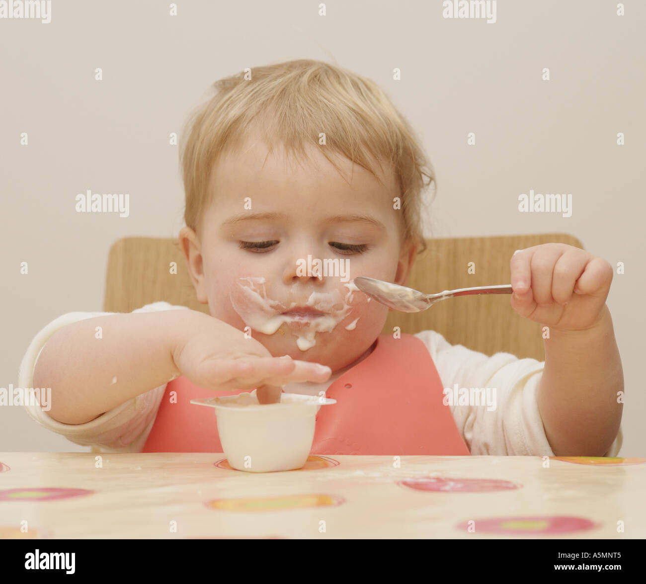 One year old child eating yogurt with a spoon and lots of food over her face Stock Photo