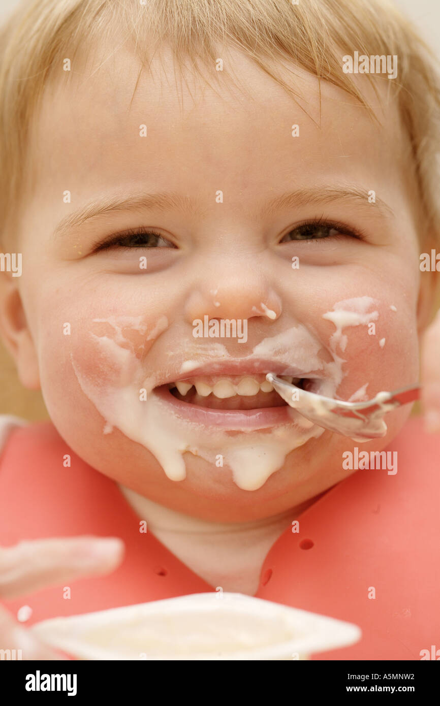 One year old child eating yogurt with a spoon and lots of food over her face Stock Photo