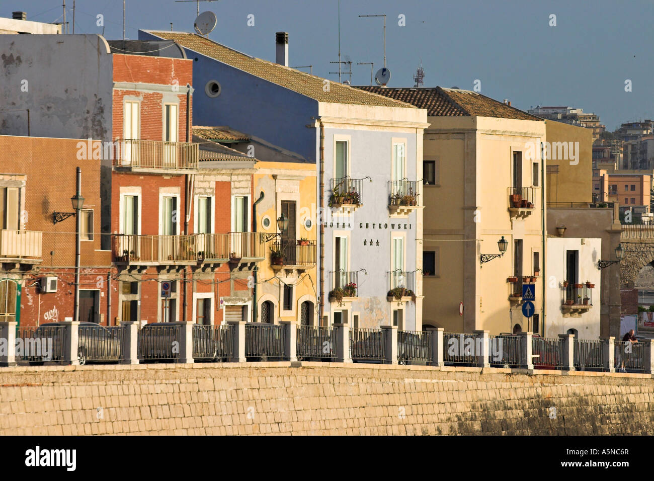 Morning Streetscape: The brightly painted houses and hotel lit by a warm rising sun face the east above the sea wall on Ortygia Stock Photo