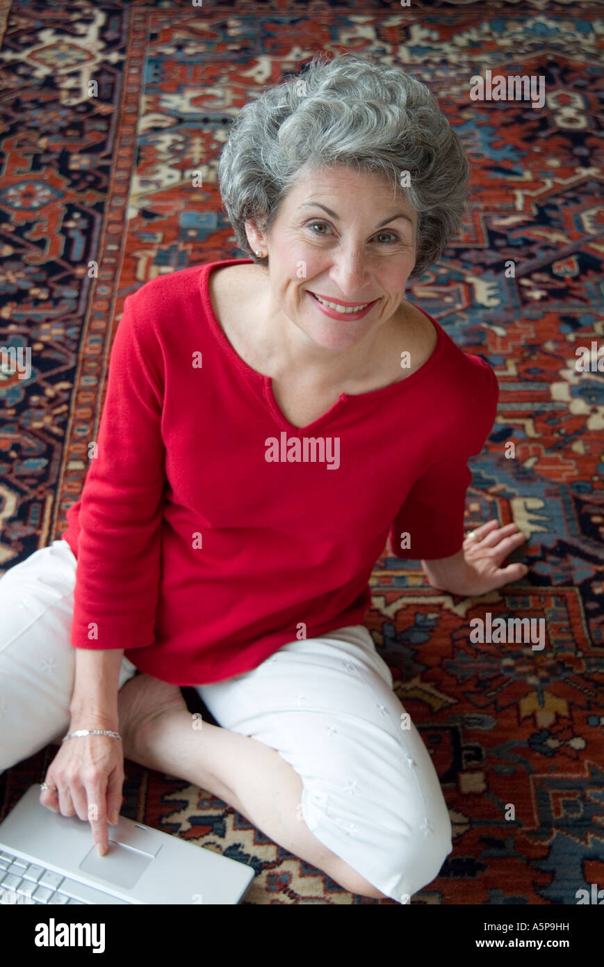 Senior woman at home with computer on carpet, smiling at camera. Stock Photo