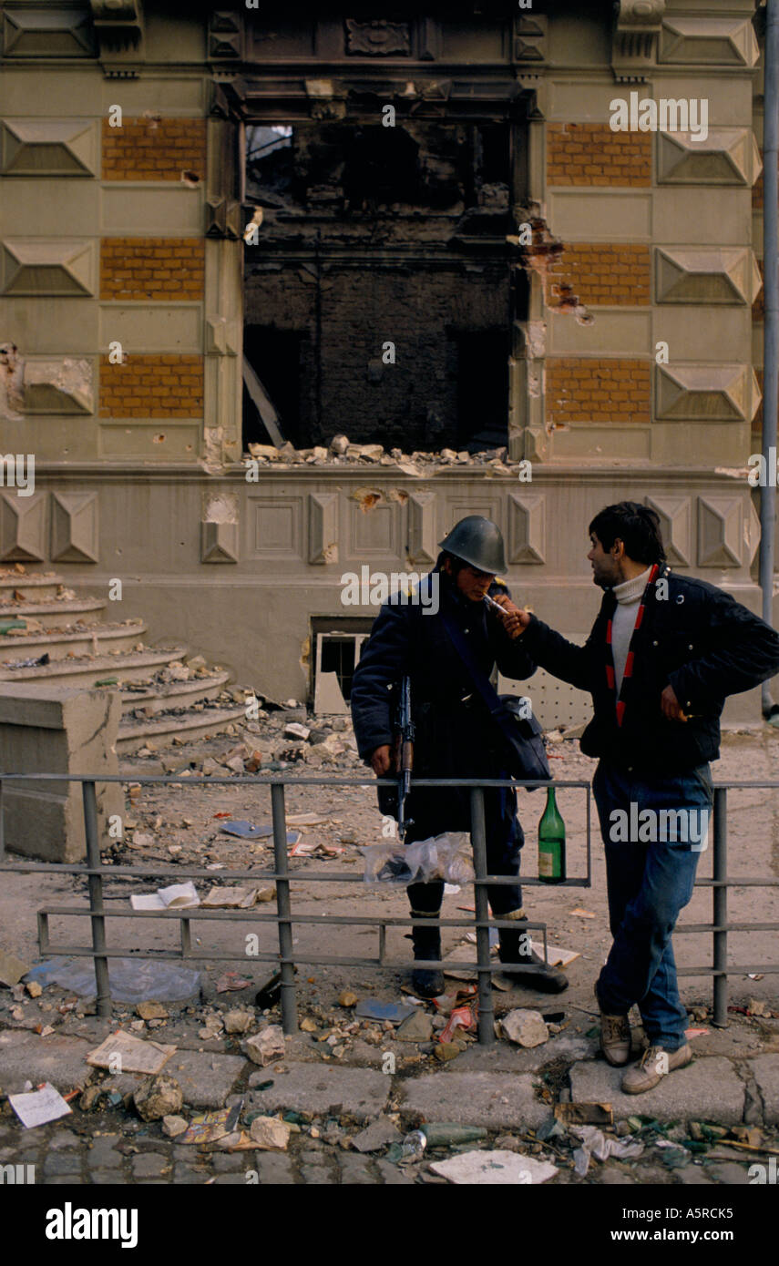 ROMANIAN REVOLUTION 1989, MAN LIGHTING A SOLDIER'S CIGARETTE AS THEY STAND  TALKING AMONGST THE RUBBLE OUTSIDE THE EMPTY SHELL OF Stock Photo