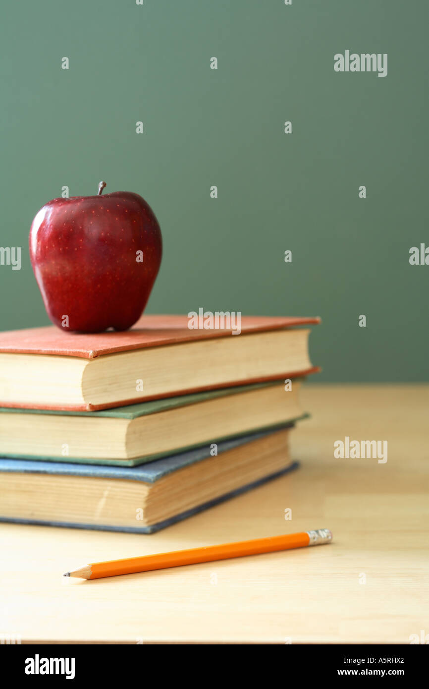 Stack of school books, red apple and pencil on desk with chalkboard in background. Stock Photo