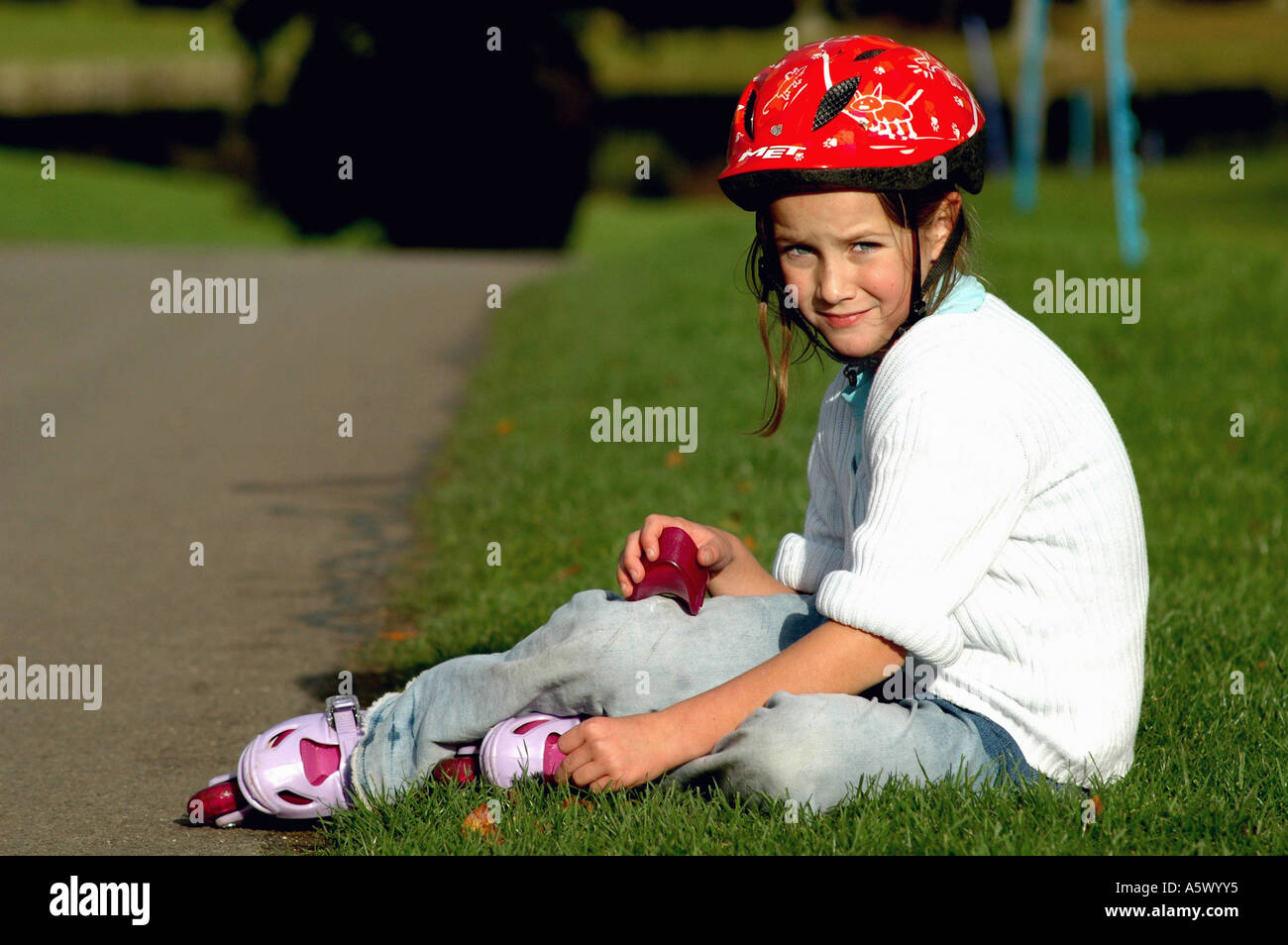young girl wearing a red safety helment and sitting on the grass in the park having a rest from rollerblading Stock Photo