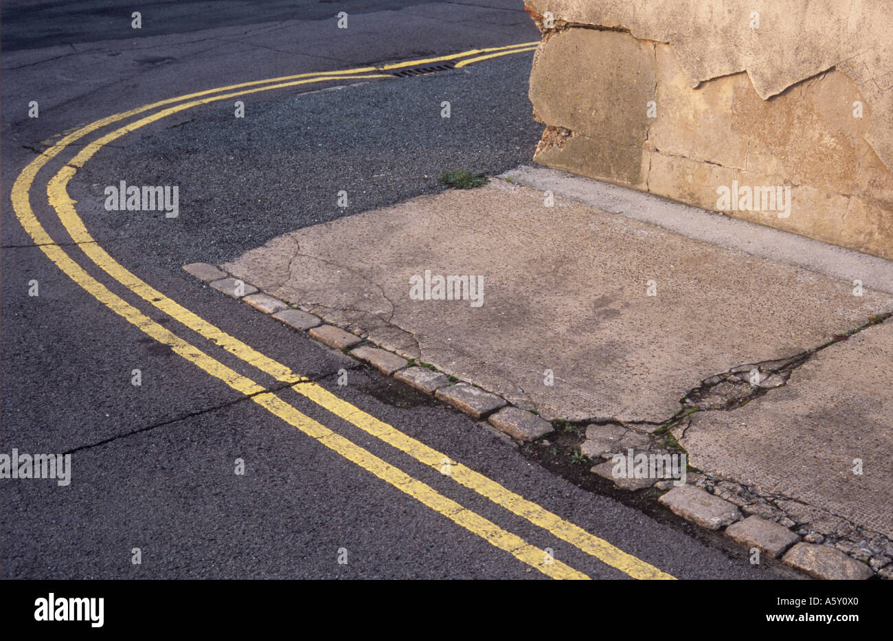 Double yellow lines curving round a street corner with disintegrating wall and helping to identify where pavement should be Stock Photo