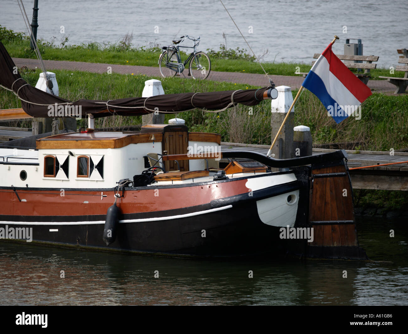 Stern of a typical Dutch flat bottom sailing ship at a dike in Woudrichem with waving Dutch flag and a bicycle parked nearby Stock Photo