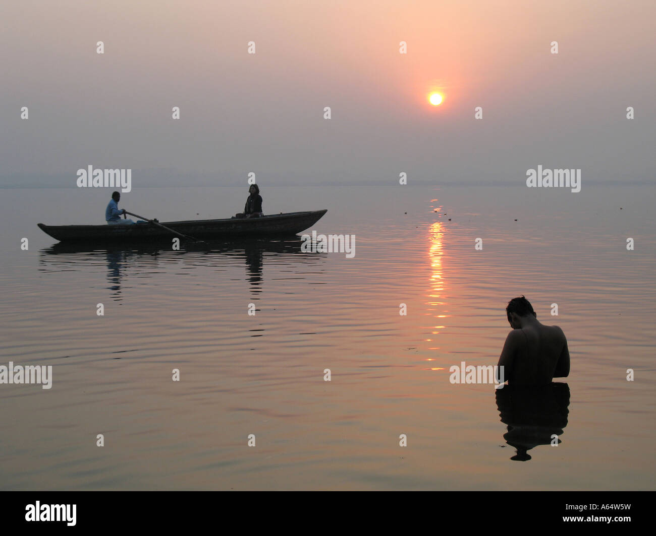 A boat passes a man praying in the river Ganges at sunrise Varanasi India Stock Photo